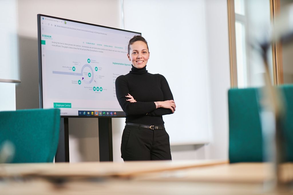 B. Renata Almeida, una empleada de Braun, de pie y sonriendo delante de un televisor que muestra gráficos en una oficina. 