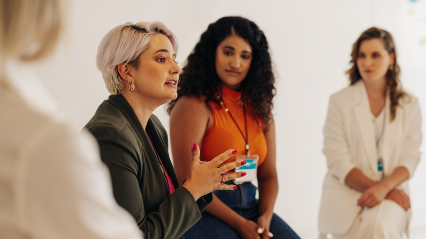 Cuatro mujeres se sientan una al lado de la otra en el programa de mentoría femenina. El objetivo es aumentar la visibilidad de las mujeres en la organización y ayudarles en su crecimiento personal. Una segunda mujer observa a la primera, una mujer de pelo gris con chaqueta verde, mientras habla y hace una señal de mano.  